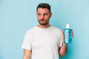 Young caucasian man holding mouthwash isolated on blue background confused, feels doubtful and...