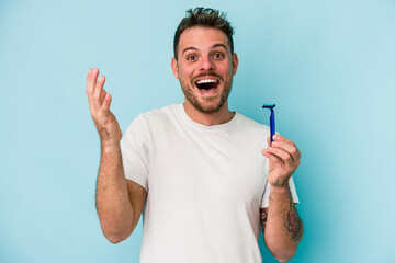 Young caucasian man shaving his beard isolated on blue background receiving a pleasant surprise,...