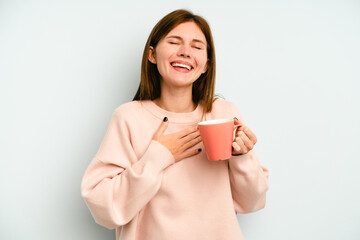 Young English woman holding a mug isolated on blue background laughs out loudly keeping hand on chest.