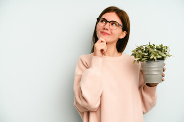 Young English woman holding a plant isolated on blue background looking sideways with doubtful and skeptical expression.