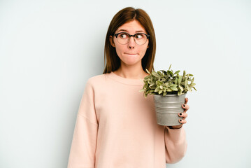 Young English woman holding a plant isolated on blue background confused, feels doubtful and unsure.