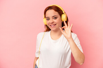 Young caucasian woman listening to music isolated on pink background cheerful and confident showing ok gesture.