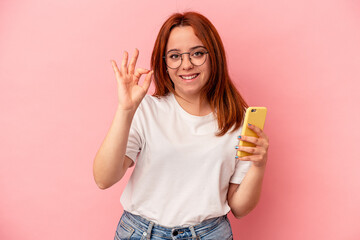 Young caucasian woman holding a mobile phone isolated on pink background cheerful and confident showing ok gesture.