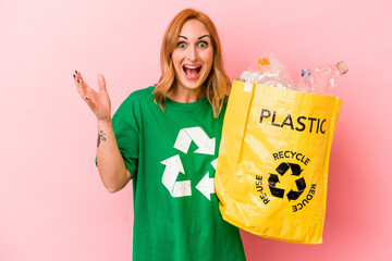Young caucasian woman recycled plastic isolated on pink background receiving a pleasant surprise,...