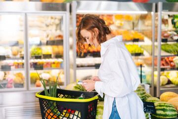 Medium shot portrait of pretty young woman checking products in grocery cart standing on background of shelves in supermarket.