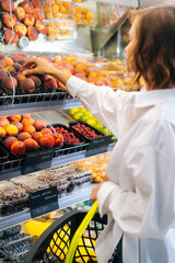 Vertical shot of young woman walking over to fruit shelf, examining fruit and putting in grocery cart. Caucasian female choosing organic foods in shop. Lady decision buying nutrition fresh food.