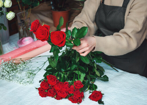 Woman florist creating a bouquet of fresh red roses. Cropped image of female florist arranging rose flowers using tools. Concept of working with flowers, floral business. Selective focus