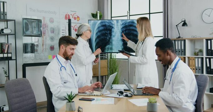 Medicine concept where attractive confident experienced multiethnic doctors working together in medical office,men typing on laptops while women discussing chest and lungs x-ray on screen