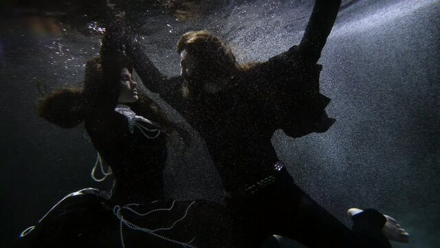 beautiful woman in dress with pearls and dramatic man in black are dancing underwater