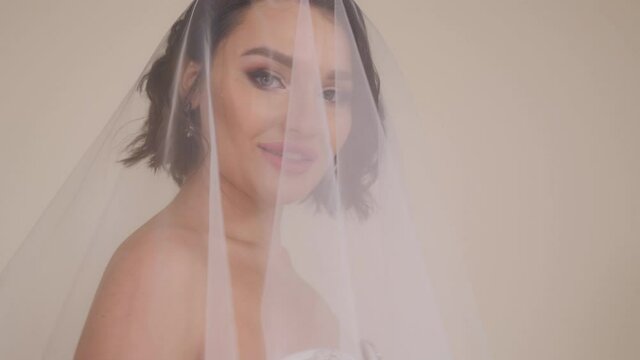 Close-up portrait of a young beautiful bride with dark hair with a head covered with a veil on a white background.