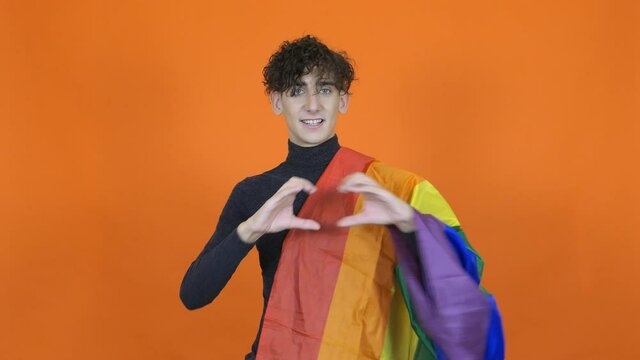 Joyful guy wrapped in a gay flag. Cute male on an orange background.