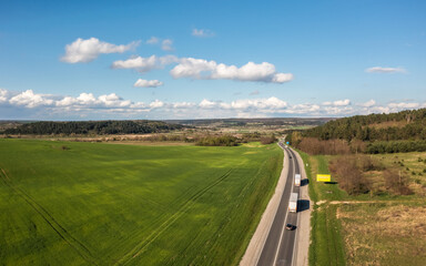 Aerial view. Green field in clear weather, the road leading into the distance. There are rare clouds in the blue sky. There is a forest on the horizon