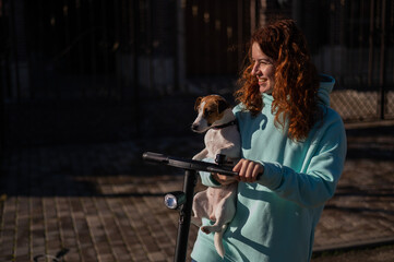 Fototapeta na wymiar A red-haired woman rides an electric scooter around the cottage village with the dog Jack Russell Terrier.