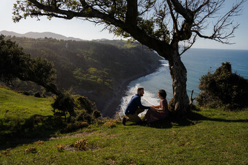 Cute young couple sitting on the grass on a cliff next to a tree