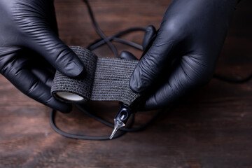 the hands of a permanent makeup artist in black sterile gloves wind a black barrier tape on a tattoo machine for protection