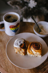 Baked cinnamon bun rolls with white vanilla bean cream frosting. Moody dark food photo. Coffee and baked pastry breakfast. Fresh baking treat. Brown cinnamon sugar. Swirls on roll. Coffee table.Brunch