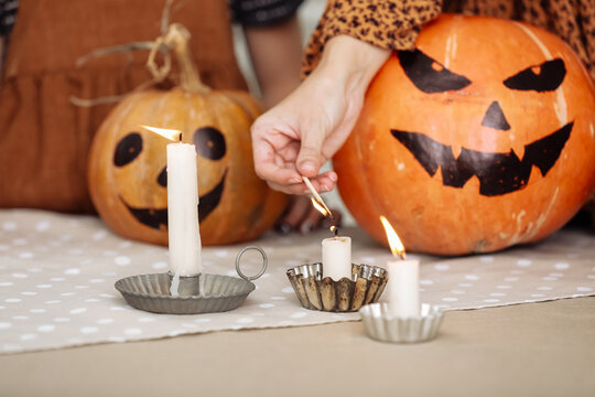 holidays and leisure concept - woman's hand with matches lighting candles at home on halloween. painted pumpkins on a background. horror theme and Hallowe'en.
