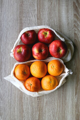 Two sustainable shopping bags filled with apples and oranges on wooden table. Top view.
