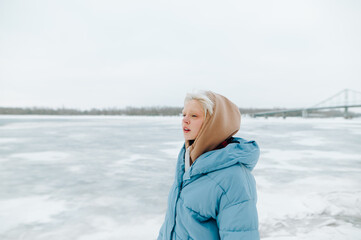 Winter portrait of a girl with blond hair on a background of frozen river and bridge, looking aside with a serious face on a beautiful winter views