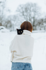 Back of a woman in a white knitted sweater with a black cat on her shoulders on a winter walk. Photography with a cat in winter.