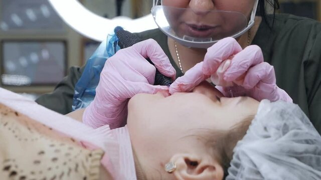 Cosmetologist wearing a mask and gloves applies permanent makeup to the lips of a young girl
