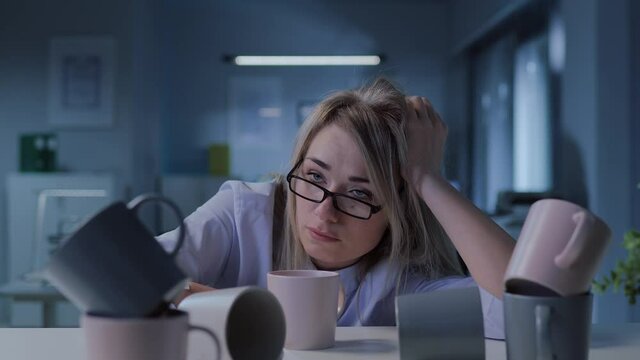 Young exhausted businesswoman sitting at desk with many empty coffee cups late in office