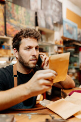 Caucasian man talks on his mobile phone with a client while checking orders and shipments in his workshop.  small business owner.