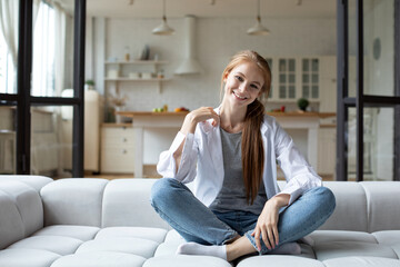 Obraz premium Portrait of happy redhead woman sitting on sofa at home, daydreaming.