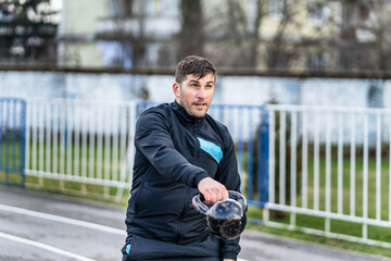 Portrait of one adult caucasian man male athlete standing outdoor at stadium track hold kettlebell in hand doing swing - strength and endurance training concept in spring or autumn day copy space