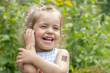 Portrait of a laughing little girl with a QR code on her shoulder against the background of nature. Concept: vaccination of children, assignment of a QR code, people with antibodies to the virus, vacc