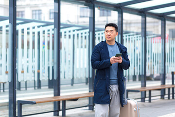 Asian businessman tourist, waiting for a taxi near the airport, uses the phone app to rent a car, a man with a big suitcase
