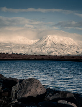Icelandic winter landscape. Mount Esja covered with snow, blue ocean waters and black rocks in foreground. No people, copyspace. 
