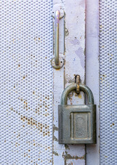 The padlock is hanging on the metal door. The lock shackle is placed in the hinges of the door. The lock is in a closed state. A metal door handle is installed above the lock.