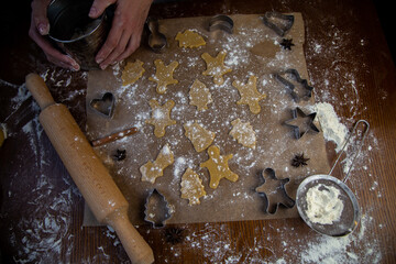 There are ready-made cookies of various shapes on the baking parchment, a rolling pin, cookie cutters and a sieve with flour are lying nearby, flour is scattered everywhere, the hands of the pastry