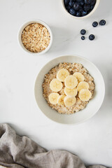 Bowl of porridge topped with banana,  a bowl of oats and a  bowl of blueberries on white marble countertop