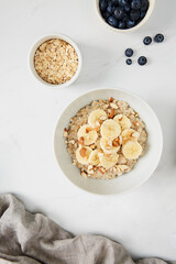 Bowl of porridge topped with banana and almonds,  a bowl of oats and a  bowl of blueberries on white marble countertop