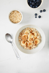 Bowl of porridge topped with banana,  almonds and cinnamon,  a bowl of oats and a  bowl of blueberries on white marble countertop