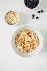 Bowl of porridge topped with banana,  almonds and cinnamon,  a bowl of oats and a  bowl of blueberries on white marble countertop