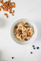 Bowl of porridge topped with banana, blueberries, almonds and cinnamon and almonds and blueberries scattered on white marble countertop