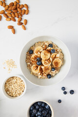 Bowl of porridge topped with banana, blueberries, almonds and cinnamon, a bowl full of oats and berries along with almonds and blueberries scattered on white marble countertop