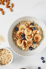 Bowl of porridge topped with banana, blueberries, almonds and cinnamon, a bowl full of oats along with almonds and blueberries scattered on white marble countertop