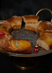 Surprise porcelain figurine of a Roscón de Reyes stands over an elegant metallic tray between two pieces of this candied cake.