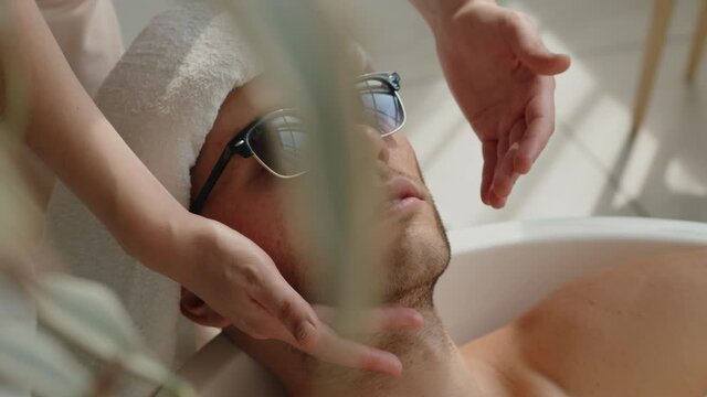 Young man in sunglasses enjoying face and head massage by woman hands in luxury spa salon ilying in bath. Sunny bright day relax in beauty resoart eco hotel. Holidays leasure recration concept