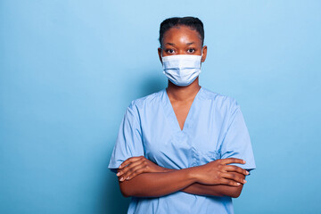 Portrait of african american practitioner nurse wearing protective medical face mask against coronavirus working at sickness treatment in studio with blue background. Health care service