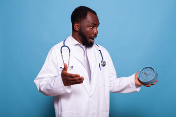 Medic in white lab coat with stethoscope holding clock feeling like out of time on blue background....