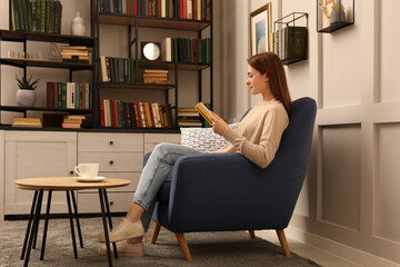 Young woman reading book in armchair indoors. Home library