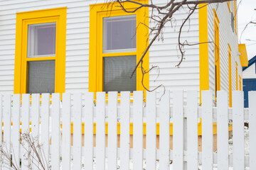The exterior of a white clapboard wooden home with two bright yellow trim double hung windows and a white wooden picket fence. A string of colored Christmas lights are strong from window to window.
