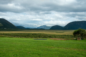 Gros Morne mountain on the Northern Peninsula of Newfoundland is flat on top with hiking trail up. The sky is cloudy with blue spots. There are hills, lush marsh, and trees in front of the mountain 