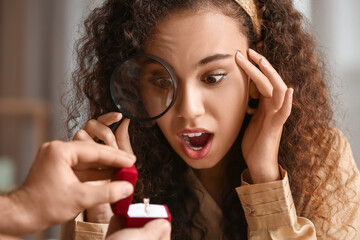 Surprised young African-American woman looking at engagement ring given by her boyfriend through magnifier at home