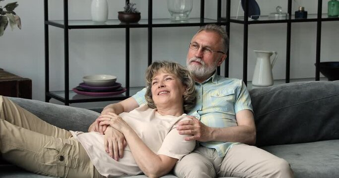 Peaceful mature couple relax on soft couch at home cuddle look at distance recall pleasant life moments in memory. Affectionate older husband sit on sofa hug serene wife from back dream of good future